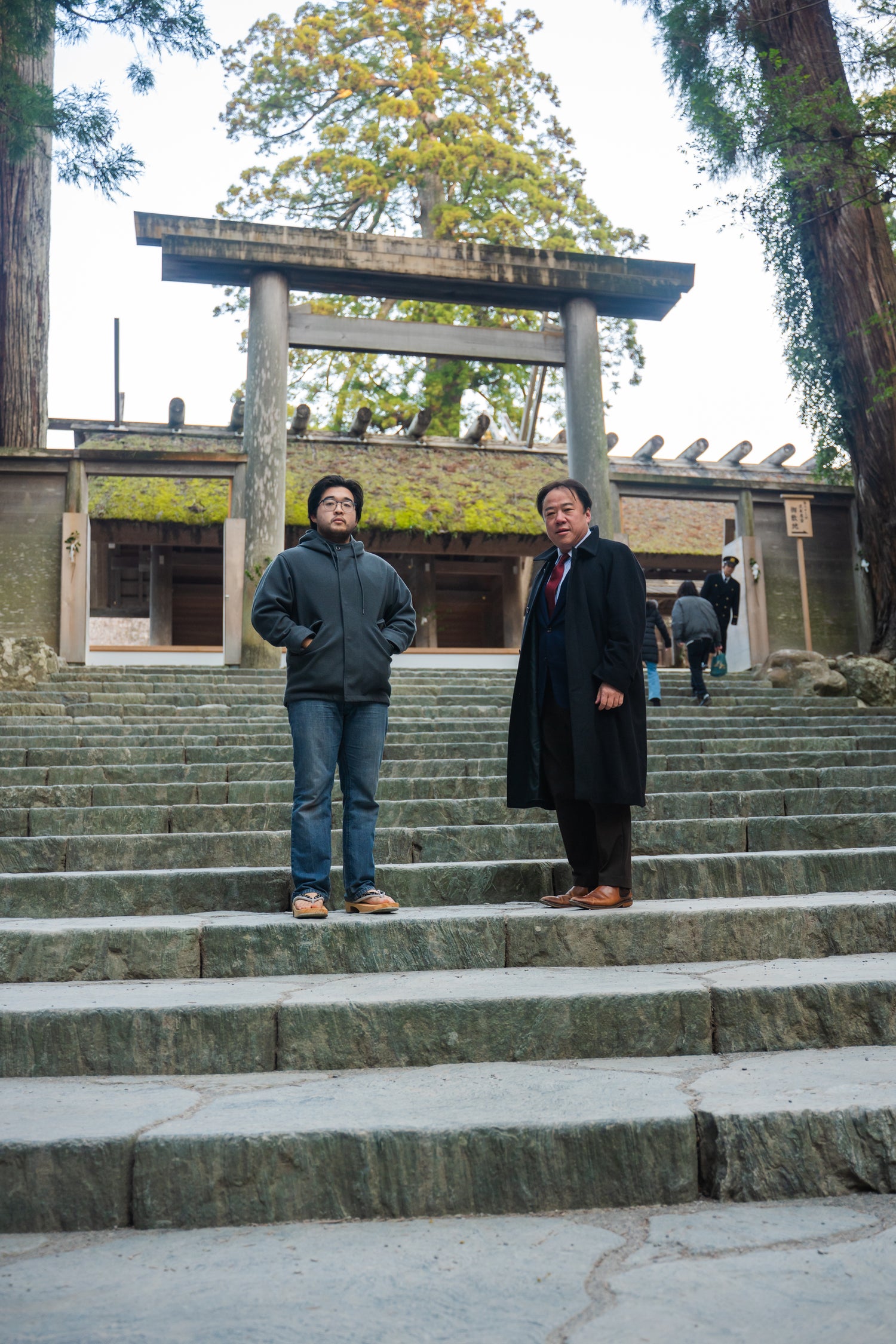 Two men standing on stone steps in front of a traditional Japanese torii gate (Ise Jingu).